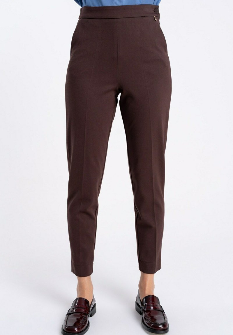 Brązowe, dopasowane do ciała spodnie sięgające kostek, noszone z ciemnoczerwonymi skórzanymi mokasynami przez stojącą osobę na jednolitym tle.