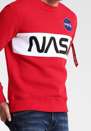 Mand iført en rød sweatshirt med et hvidt panel og sort NASA-tekst, en NASA-logo-patch og en rød "Remove Before Flight"-mærkat på ærmet.