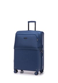 Valise rigide bleue avec un design texturé à côtes, poignée rétractable, quatre roues et une poche zippée à l'avant. Étiquette de marque noire visible.