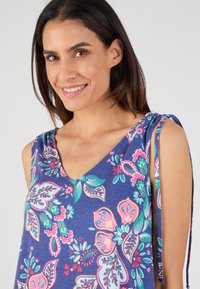 Top sans manches en V en tissu bleu avec un motif floral coloré ; se caractérise par des détails de lien ajustables sur les épaules.