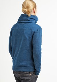 Pullover de forro polar azul y texturizado con un cuello alto y doblado y mangas largas; cuenta con un dobladillo ajustado y un material liso y elástico.