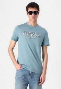 T-shirt azzurro chiaro a maniche corte con la scritta "GUESS EST. 1981" stampata in colori a contrasto; tessuto morbido; abbinato a jeans in denim chiaro e occhiali da sole.