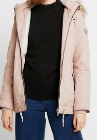 Jasnoróżowa puchowa kurtka z zamkiem i kapturem obszytym futrem, z czarnym, prążkowanym swetrem pod spodem oraz niebieskimi dżinsami.