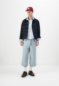 Donkerdenim jack over een lichtblauw gestreept shirt, wit T-shirt, brede lichtblauwe spijkerbroek en bruine schoenen met witte sokken. Rode pet.