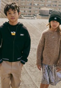 Zwarte zip-up hoodie, kaki cargobroek met ritszakken. Beige chunky gebreide trui, metalen zilveren paillettenrok, zwarte beanie.