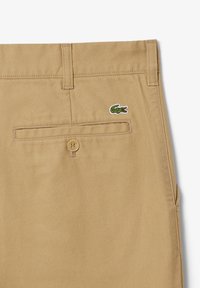 Bermudas de algodón beige con un bolsillo trasero, que cuentan con un cierre de botón y un pequeño logo de cocodrilo verde bordado en la tela.