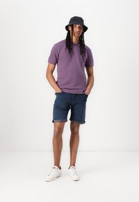 Ljubičasta polo majica, tamne traper šortse s preokrenutim rubovima, bijele tenisice i crna kapa u obliku kante. Jednostavan, ležeran outfit, minimalni uzorci.