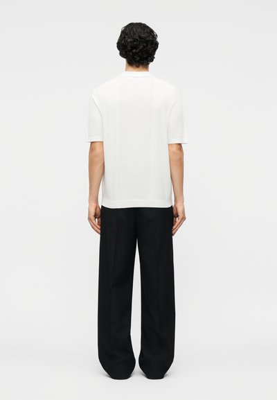 Άνδρας με σγουρά μαλλιά που φοράει λευκό πουκάμισο με κοντά μανίκια και υφασμένη πίσω πλάτη, καθώς και χαλαρό μαύρο παντελόνι με φαρδιά μπατζάκια, στέκεται με την πλάτη προς τα εμπρός.