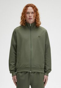 Giacca a zip verde oliva con alto colletto, polsini e orlo a costine. Presenta un piccolo logo sul petto e tasche laterali. Texture liscia.