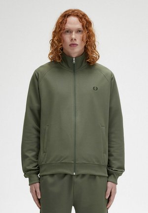 Giacca a zip verde oliva con alto colletto, polsini e orlo a costine. Presenta un piccolo logo sul petto e tasche laterali. Texture liscia.