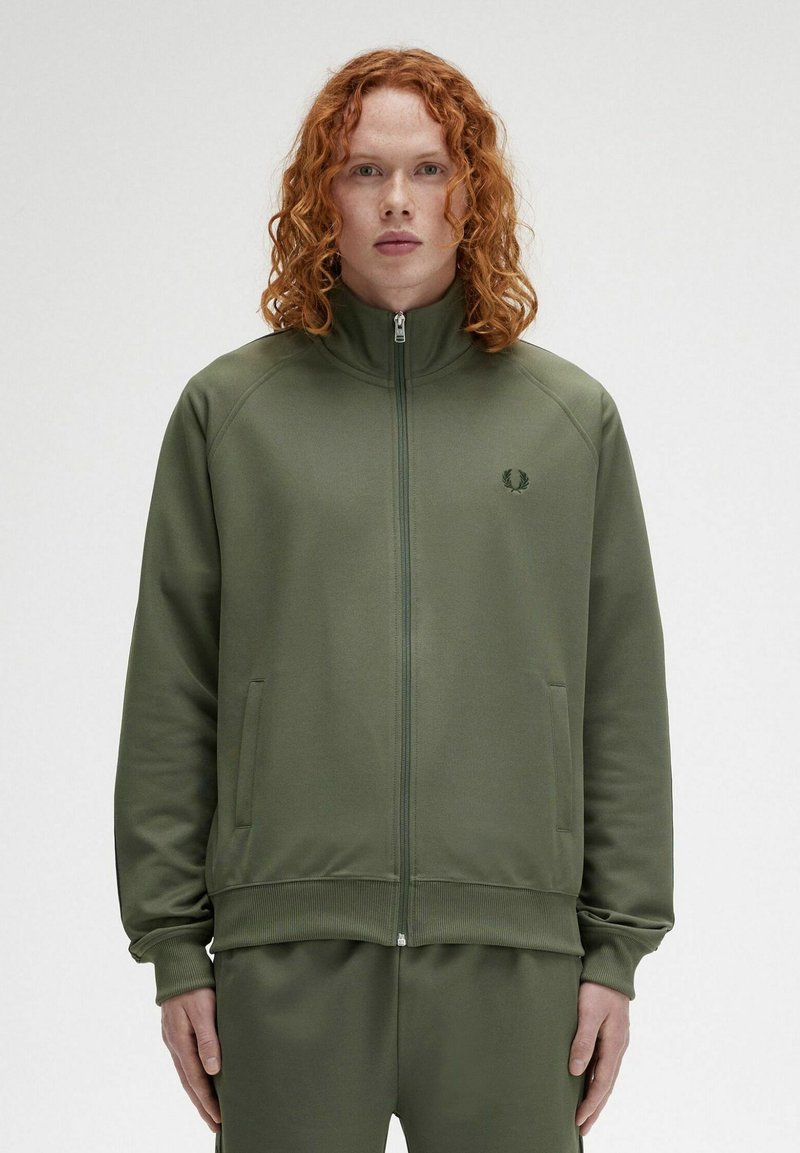 Giacca a zip verde oliva con alto colletto, polsini e orlo a costine. Presenta un piccolo logo sul petto e tasche laterali. Texture liscia.