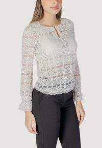 Blusa di pizzo color crema chiaro con motivo floreale, scollo rotondo, dettaglio a chiave e maniche arricciate, abbinata a pantaloni neri aderenti.