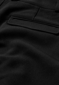 Tissu en pantalon noir avec une texture lisse, comportant une poche arrière unique et des détails de couture discrets. Le matériau semble être tissé.