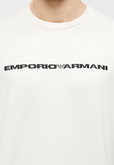 Άτομο που φοράει λευκό μπλουζάκι με στρογγυλή λαιμόκοψη και έντονη μαύρη επιγραφή «EMPORIO ARMANI» με λογότυπο στο κέντρο του στήθους.
