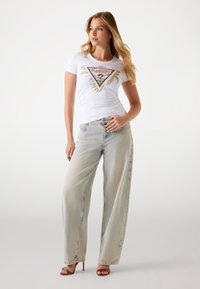 Weißes kurzärmliges Hemd mit einem bunten, dreieckigen Grafikdesign, kombiniert mit hellblauen weit geschnittenen Jeans. Absatzsandalen runden den Look ab.