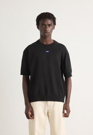 Black short-sleeve knit sweater with ribbed neckline and hem, featuring a small blue logo patch on the chest. Worn with light-colored pants.