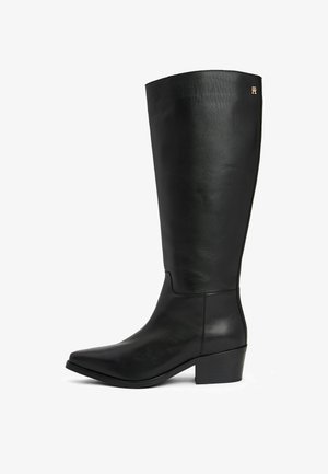 Botte noire en cuir montant jusqu'au genou avec un petit talon bloc et un petit logo doré près du bord supérieur, bout anglé sur fond blanc.
