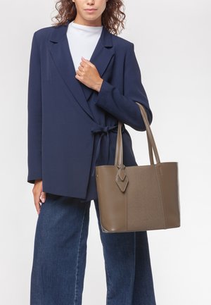 Borsa a tracolla marrone con finitura testurizzata, doppi manici e un'etichetta decorativa. Indossata con un blazer blu navy e jeans a gamba larga.