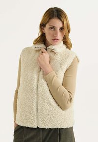 Chaleco de forro polar color crema con un collar alto, cremallera larga y un parche circular en el pecho. Tejido suave y texturizado.