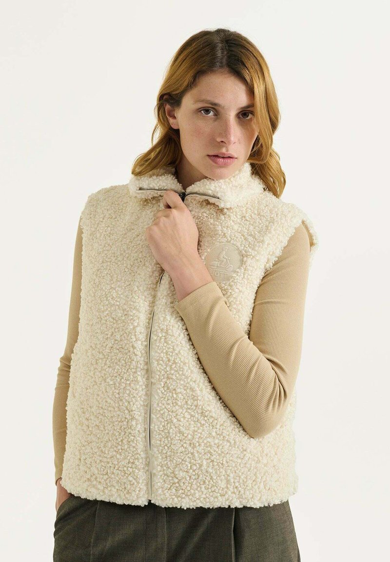 Chaleco de forro polar color crema con un collar alto, cremallera larga y un parche circular en el pecho. Tejido suave y texturizado.