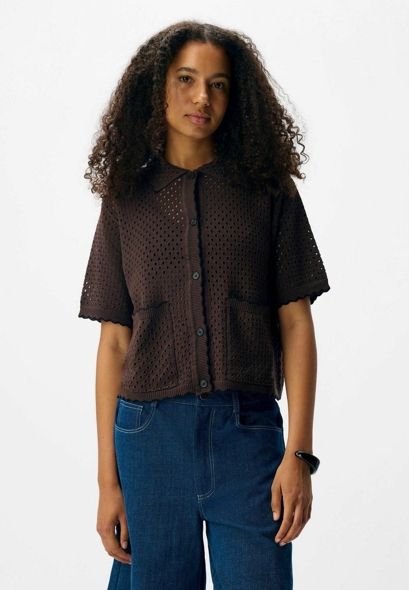 Vrouw met krullend haar die een donkerbruine oogjes knoop-blouse met korte mouwen en een blauwe spijkerbroek draagt, staande tegen een effen witte achtergrond.