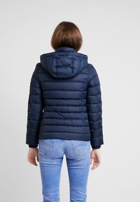 Tmavě modrá péřová bunda s kapucí, s vodorovnými prošívanými panely a pletenými manžetami. Pohled ze zad ukazuje přiléhavý střih a hladkou texturu.