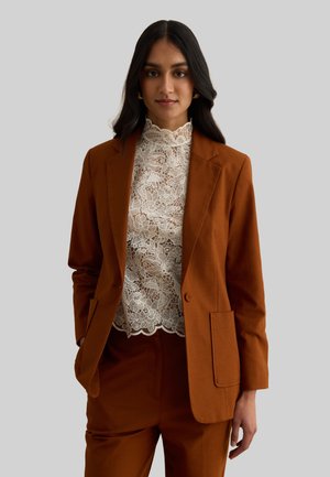 Mujer vestida con un traje color óxido y una blusa blanca de encaje floral con cuello alto, de pie frente a un fondo gris liso.