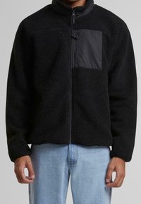 Czarna polarowa kurtka z zapięciem na zamek z przodu oraz matową czarną kieszenią na piersi. Posiada teksturowaną powierzchnię i luźny krój. Noszona na jasnoniebieskich dżinsach.