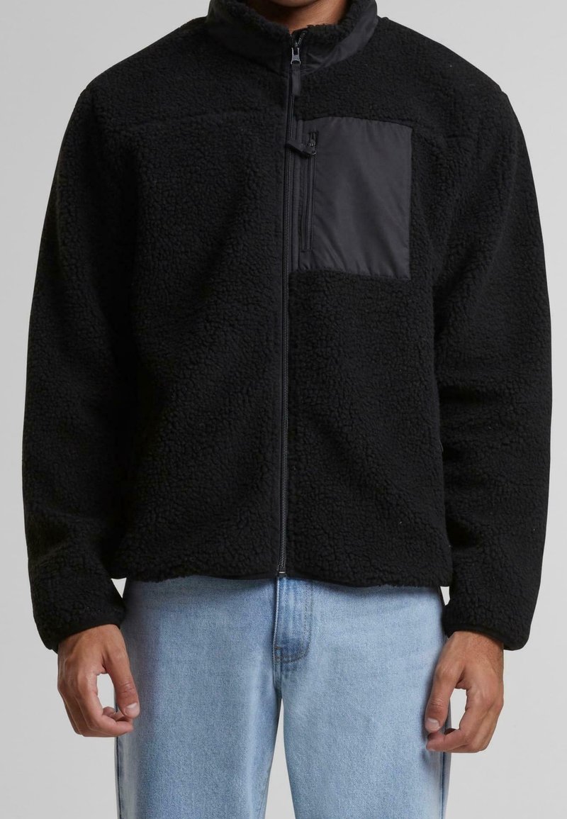 Schwarze Fleecejacke mit einem Reißverschluss vorne und einer mattschwarzen Brusttasche. Sie verfügt über eine strukturierte Oberfläche und einen lockeren Schnitt. Getragen über hellblauen Jeans.