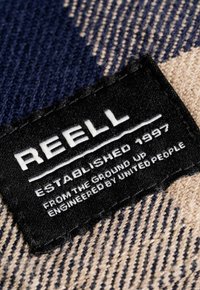 Etiqueta de tela negra con texto blanco que dice "REELL," "ESTABLECIDA EN 1997," y detalles adicionales. El fondo presenta un patrón tejido en beige y azul.