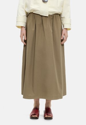 Osoba ubrana w beżową długą spódnicę, kremową kurtkę i czerwone chodaki z odkrytymi palcami stojąca na jednolitym białym tle.