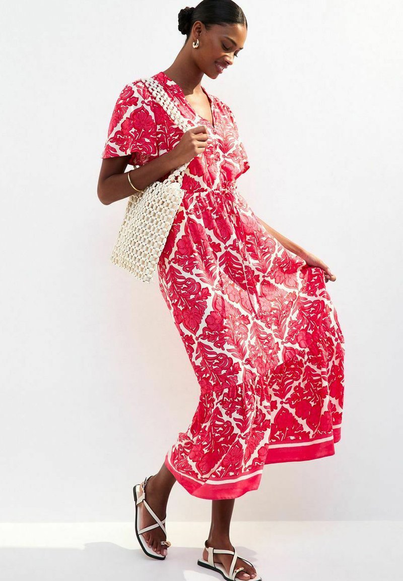 Robe maxi rouge à fleurs avec un décolleté en V, des manches courtes et une taille froncée. Associée à un sac tissé blanc et des sandales plates.