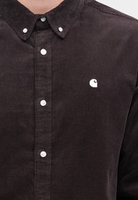 Σκούρο καφέ πουκάμισο από κορντούρα με γιακά που κλείνει με κουμπιά, διαθέτει λευκά κουμπιά και ένα μικρό λευκό λογότυπο στο αριστερό στήθος.