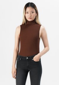 Jonge vrouw met lang haar draagt een mouwloze bruine coltrui en zwarte jeans, poseert met één hand in haar zak tegen een witte achtergrond.