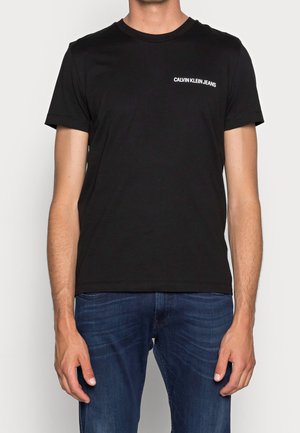 Mand iført sort T-shirt med korte ærmer med hvid "Calvin Klein Jeans" tekst på brystet og blå denim jeans, stående mod en ensfarvet baggrund.