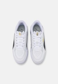 Białe syntetyczne sneakersy z siateczkowymi akcentami, płaskimi sznurowadłami, perforowanym przodem oraz czarnym paskiem po boku; mają żółtą metkę z logo.