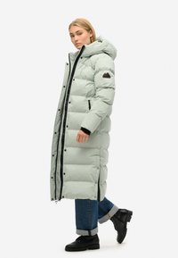 Lys grøn lang pufferjakke med hætte, der har en sort lynlås, sidelommer og et bjerglogo patch på venstre arm. Båret med sorte støvler.