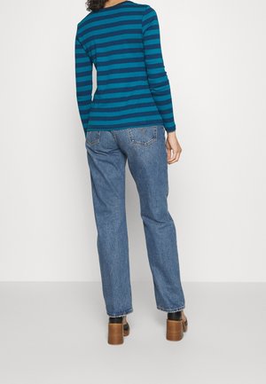 Person iført blå og turkisstribet langærmet skjorte, blå højtaljede jeans og brune sko med hæl, der står med ryggen til.