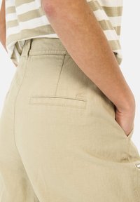Beige Baumwollhose mit entspannter Passform, ausgestattet mit einer Rückentasche mit rechteckigem Klappverschluss und subtilen Nähdetails. Gestreiftes Hemd oben sichtbar.