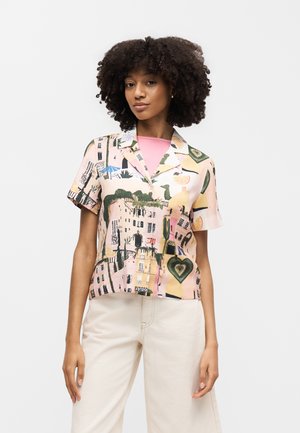 Žena s kučeravými vlasmi nosiaca krátky rukáv, košeľu na gombíky s farebnými vzormi mestských scenérií a srdiečok, skombinovanú so svetlo béžovými nohavicami.