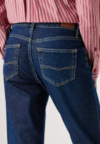 Σκούρα μπλε τζιν από denim με λεία υφή, με αντίθετη κίτρινη ραφή και δύο πίσω τσέπες, σχεδιασμένα με ψηλή μέση.