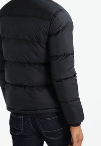 Black puffer jacket with horizontal quilted sections, made of lightweight, shiny material. Features a rounded collar and long sleeves.