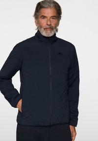 Laivastonsininen puffer-takki, jossa on vetoketjullinen etuosa, korkea kaulus ja sivutaskut. Takissa on hienovarainen logo rinnassa ja sileä, matta pinta.