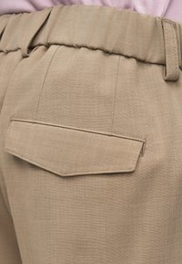 Calças bege com tecido texturizado, apresentando um bolso traseiro único com aba pontiaguda. Cintura elástica para ajuste confortável.