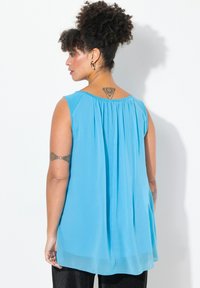 En blå, ärmlös topp i lättviktig tyg med veck längs ryggen för extra textur och flöde. Nederkant sitter vid höfterna.