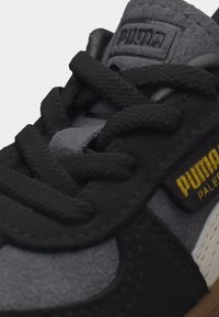 Κοντινό πλάνο μαύρου και γκρι αθλητικού παπουτσιού Puma με μαύρα κορδόνια και κίτρινη ένδειξη «Puma Pale» στο πλάι και ανάγλυφο λογότυπο Puma στη γλώσσα.