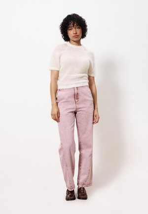 Jean taille haute rose clair avec un aspect délavé, associé à un pull léger à manches courtes blanc. Le modèle se tient sur un fond uni.
