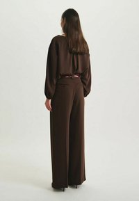Blouse en satin marron à épaules décontractées, associée à un pantalon marron ample avec une finition polie et une ceinture accentuée de rouge.