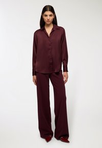 Burgundowa satynowa koszula z guzikami z kołnierzykiem, długimi rękawami i luźnym krojem, w połączeniu z dopasowanymi spodniami o szerokich nogawkach oraz czerwonymi, szpiczastymi butami.