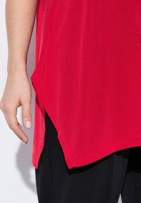 Maglietta rossa con una texture liscia, presenta un orlo curvo e spacchi laterali, abbinata a pantaloni neri. Si concentra sul colore vivace e sul taglio.